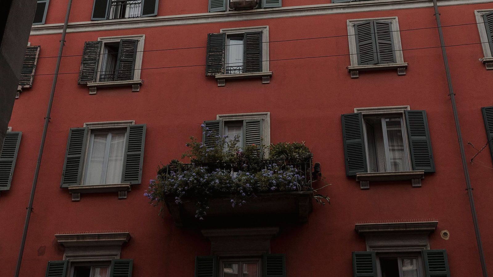 Facciata rossa di un palazzo milanese con persiane verdi e un balcone fiorito